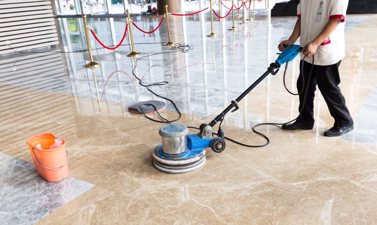 A professional cleaner operates a blue floor buffer on a shiny, reflective marble lobby floor.