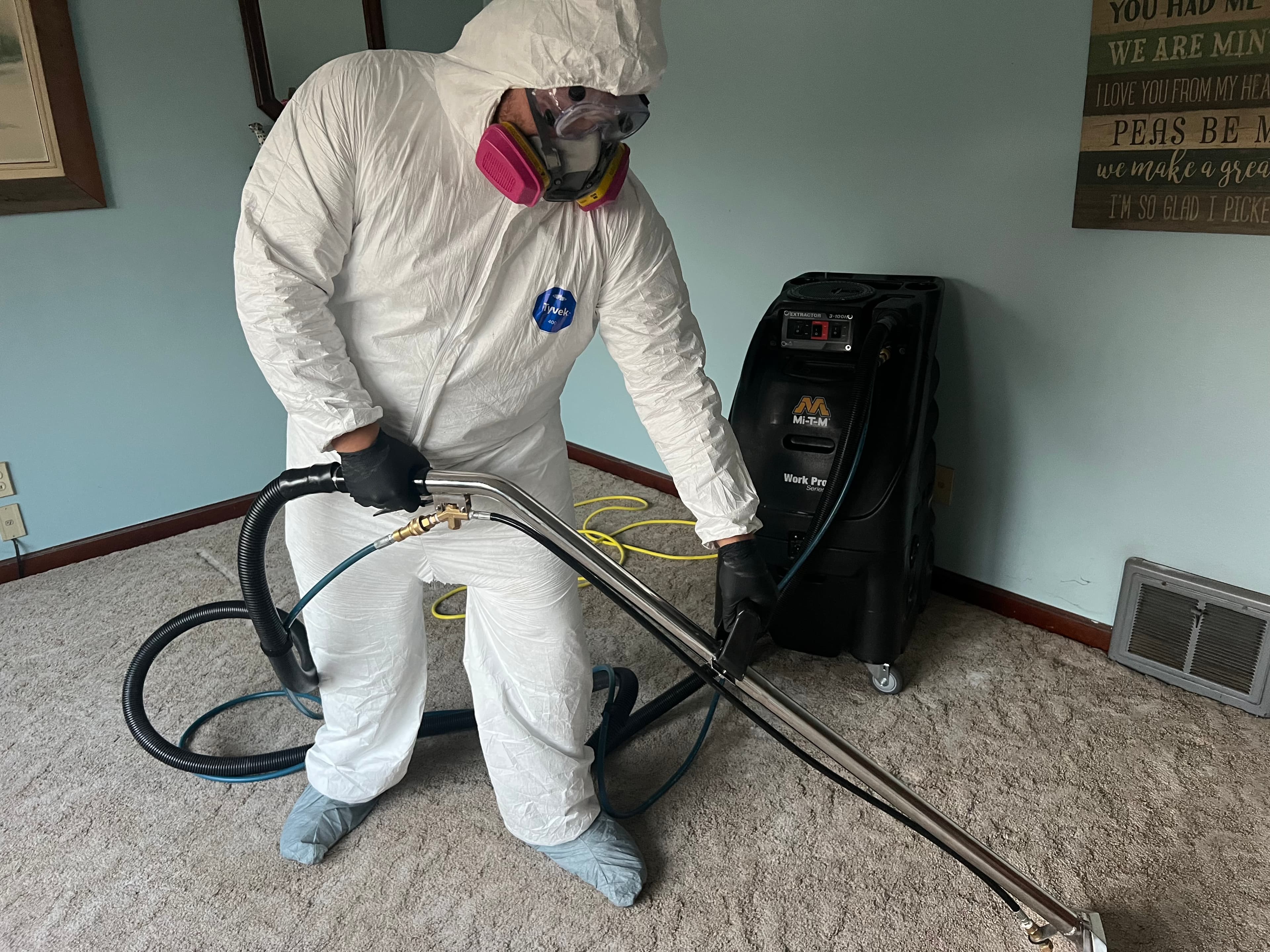 Professional in a white hazmat suit and respirator deep cleans carpet with an extractor.