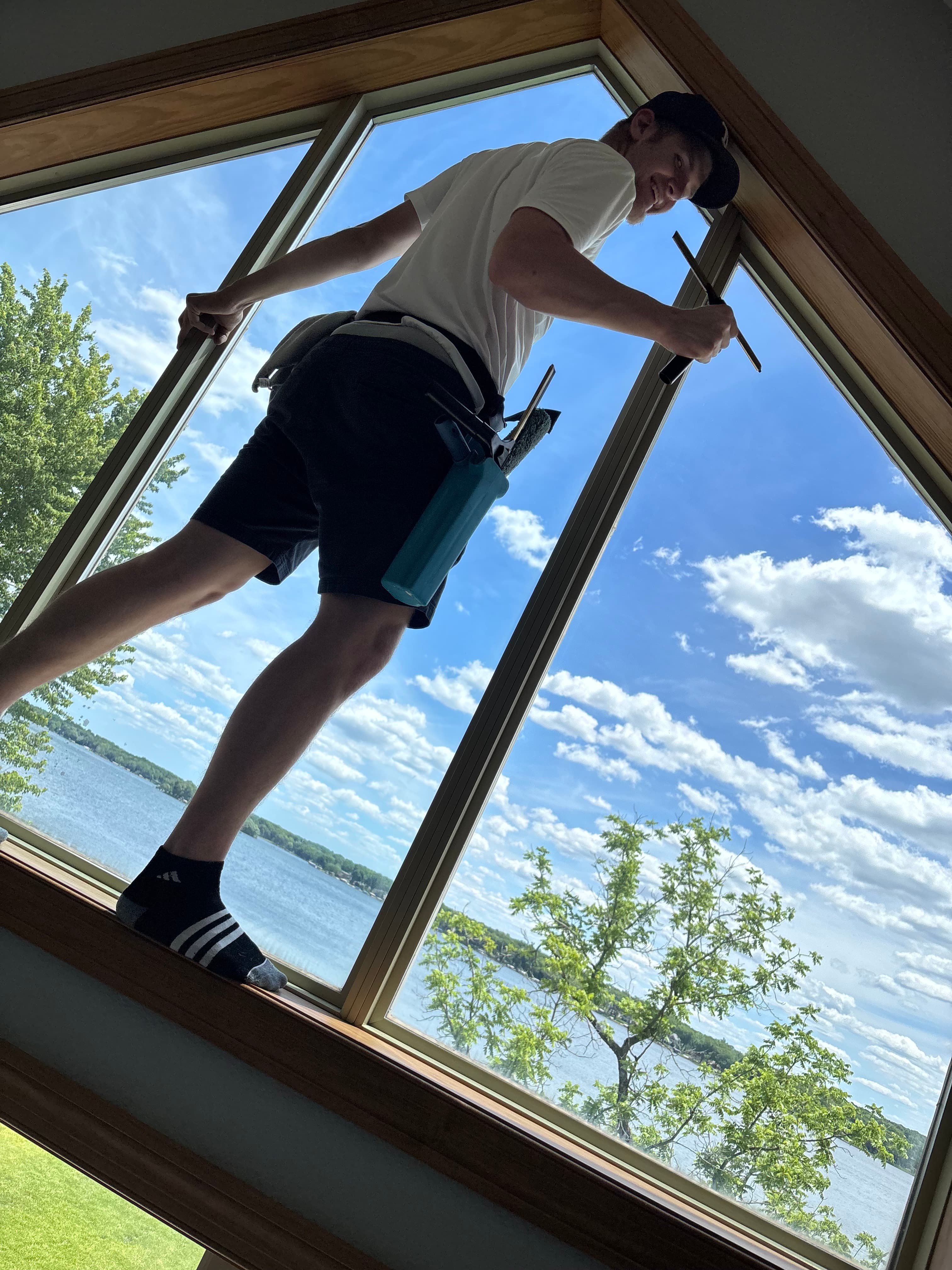 Man cleaning high windows on a ledge overlooking a scenic lake under a blue sky.