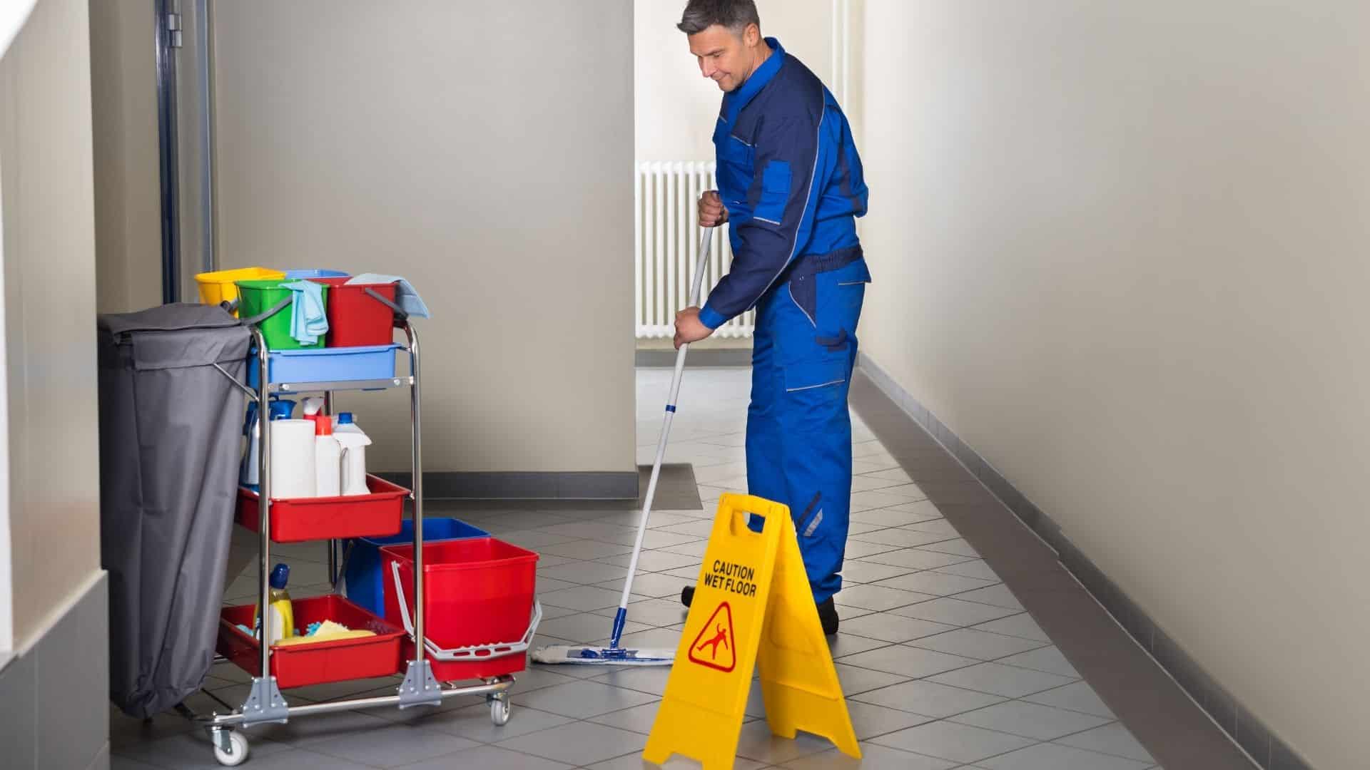 Janitor in blue uniform mopping a tiled floor next to a yellow wet floor sign.